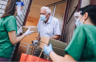 health care workers in scrubs, surgical masks and face shields stand on the porch of an elderly man, also masked. they are delivering boxes of medical supplies.