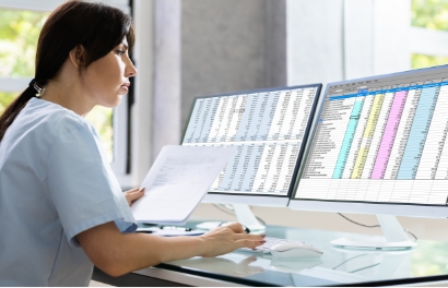 a woman in scrubs inputs data from paperwork into a computer displaying large, colorful spreadsheets on two screens