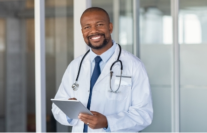 a Black doctor in labcoat and stethoscope holds a clipboard while smiling toward the camera