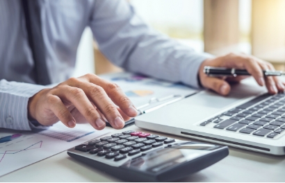 hands of a person dressed in a blue button down shirt as they use a calculator and a laptop