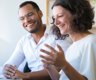 a man and woman sitting next to each other smiling and gesturing, mid-conversation