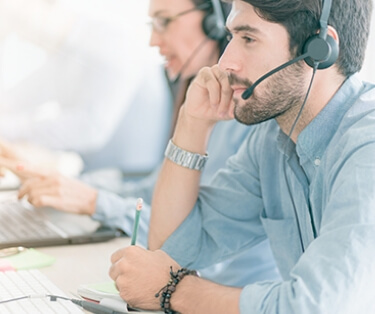 a row of people in blue button down shirts. they are talking on headsets while using computers.