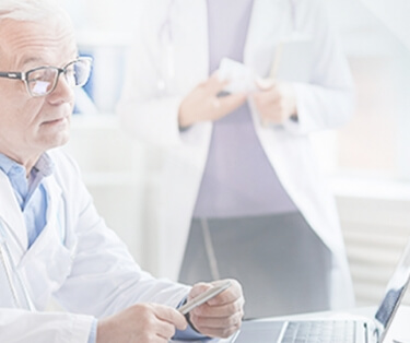 a white haired doctor wearing glasses, lab coat and stethoscope sits at a desk looking at a laptop screen